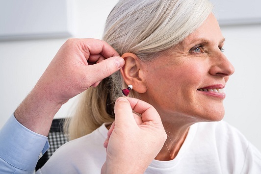 Ältere Frau bekommt modernes Hörgerät ins Ohr eingesetzt, Hörakustiker berät freundlich.